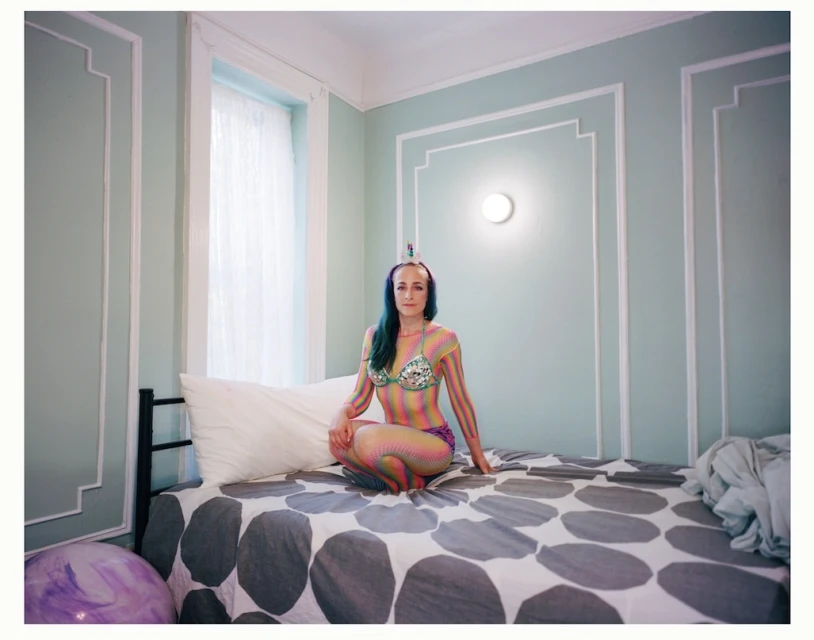 Adult female with rainbow body paint and unicorn horn sitting on a bed in an indoor bedroom with light green walls and minimal decor.