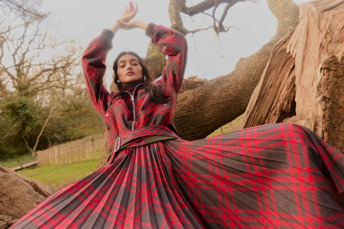 Woman in a red and black plaid pleated skirt and matching jacket posing in front of a large old tree.