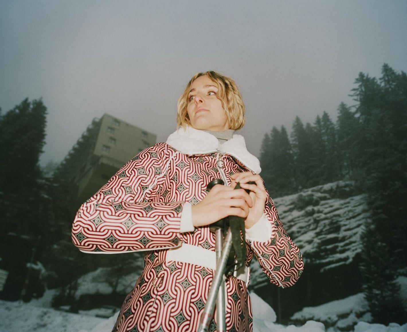 Woman in patterned coat with ski poles poses outdoors on snow-covered ground with background of evergreens and a distant building.