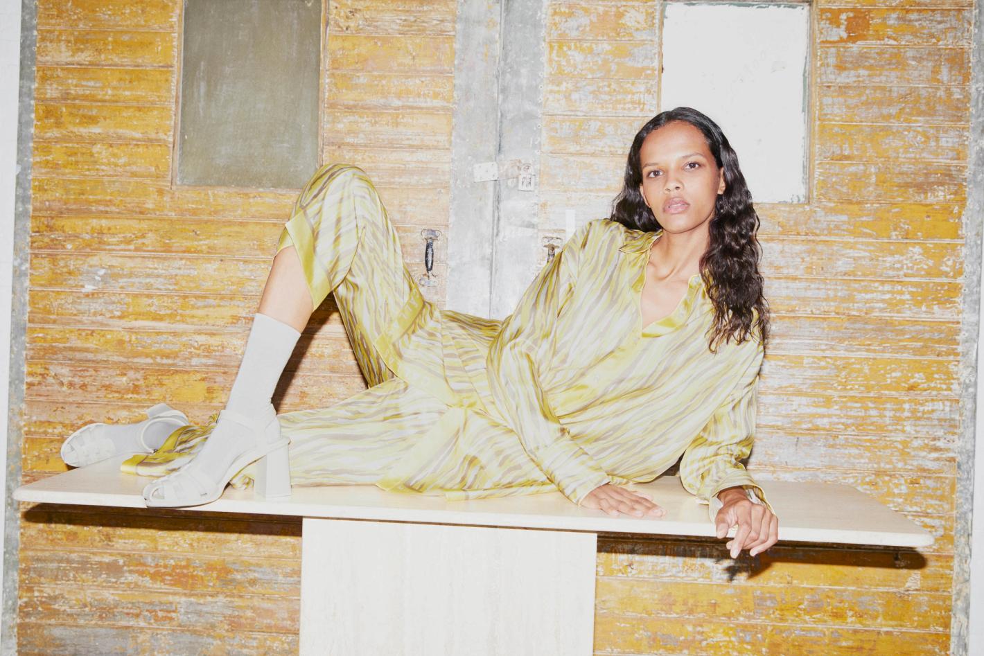 Elegant woman poses in a yellow striped loungewear set, complemented by white heeled sandals and socks, against a textured backdrop.