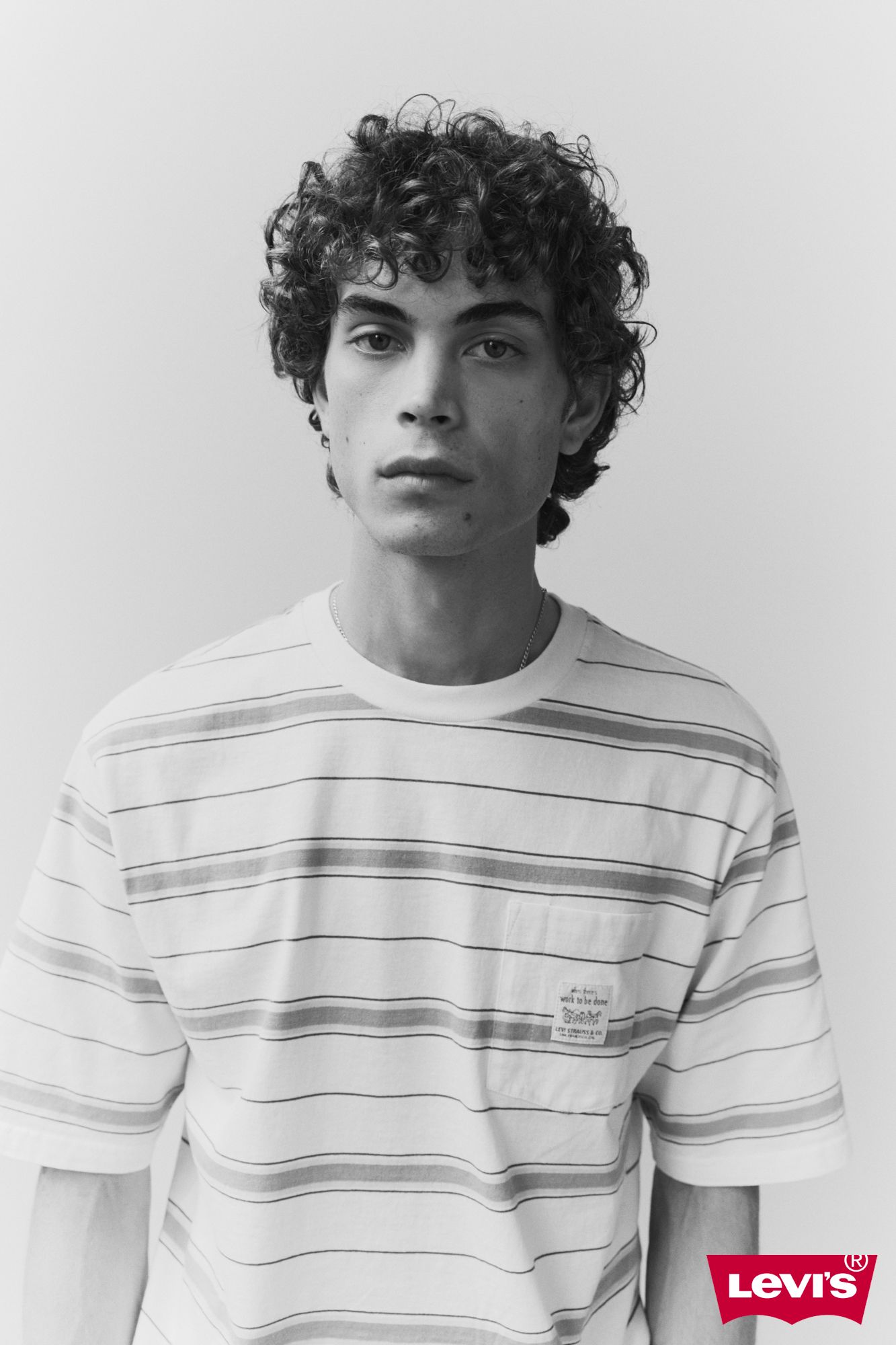 Portrait of a young adult man in a striped t-shirt. Neutral facial expression, curly hair, studio lighting, and white background.