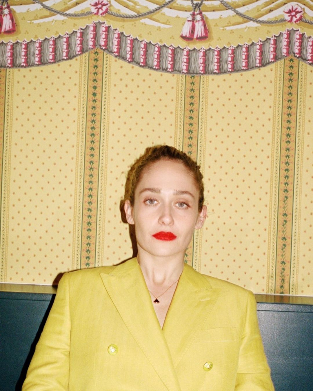 Adult woman in yellow suit poses in front of a vintage wallpaper backdrop. She has red lipstick and her hair is pulled back.