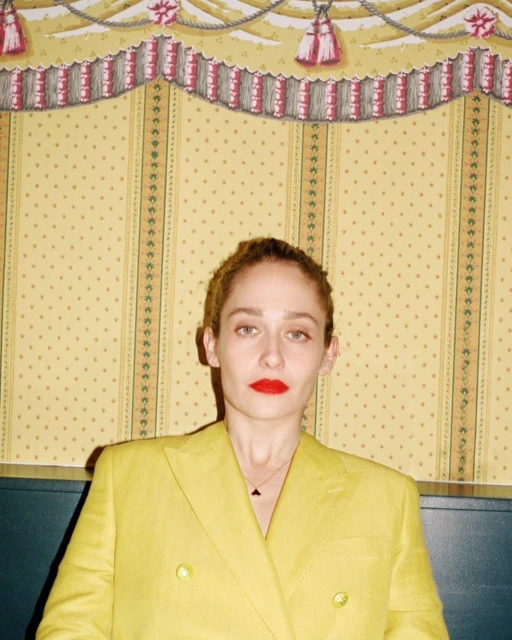Adult woman in yellow suit poses in front of a vintage wallpaper backdrop. She has red lipstick and her hair is pulled back.