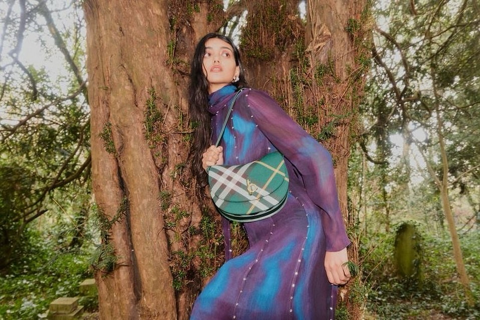 Fashion shot of a woman in a purple dress and green plaid purse posing next to a tree in a natural outdoor setting.