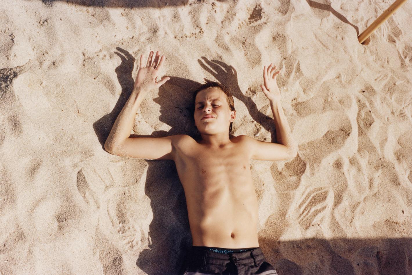 Child lying on a sandy beach with arms raised, bathed in sunlight, wearing dark shorts, capturing a relaxed vacation moment.