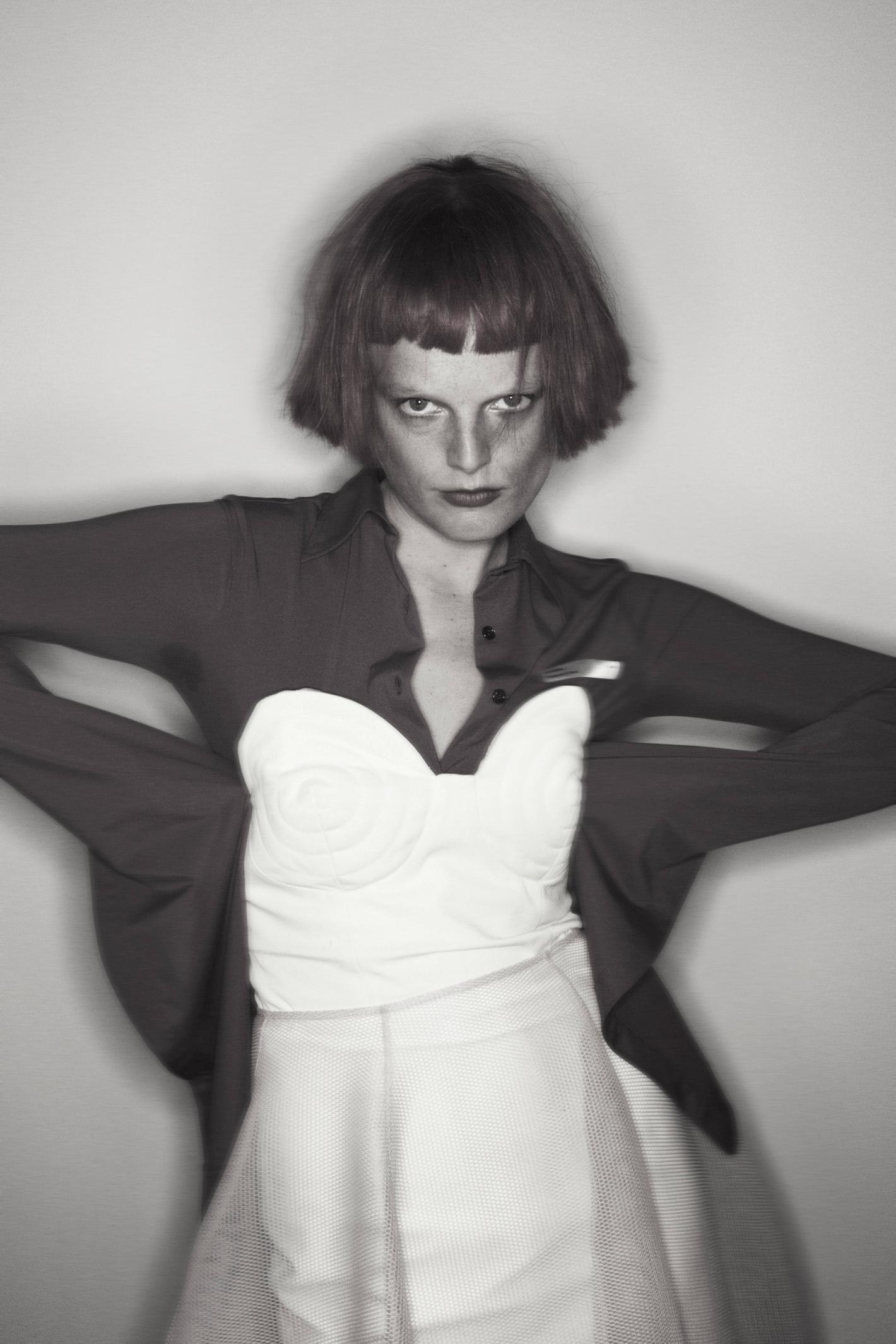 Monochrome portrait of woman with short hair wearing a red shirt over a white corset top and white skirt against a neutral backdrop.
