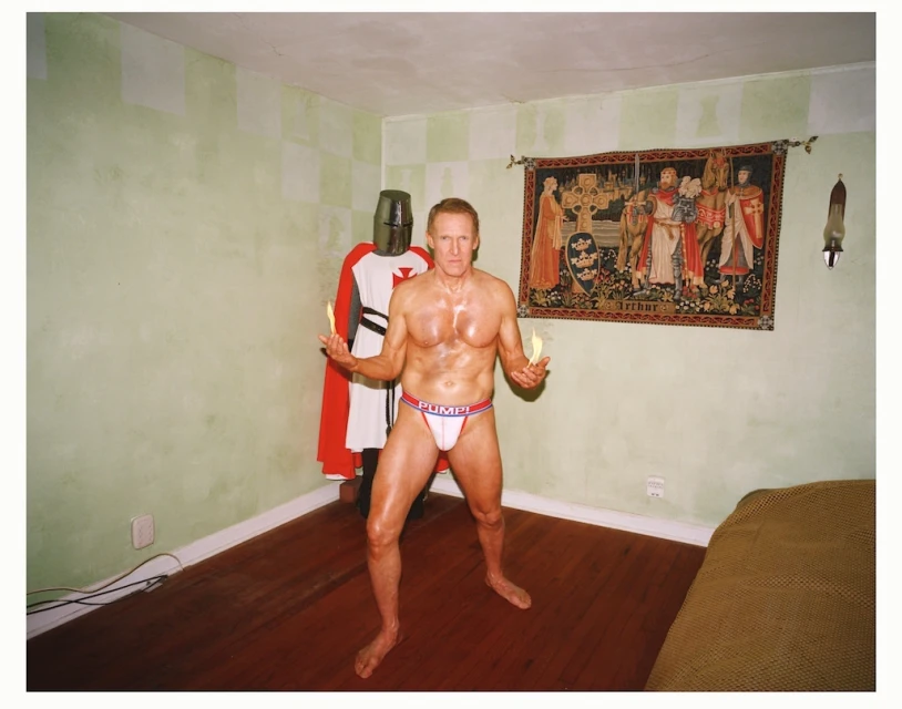 A man stands holding lit candles in underwear, with a knight in armor behind, in an indoor setting with a tapestry on the wall.