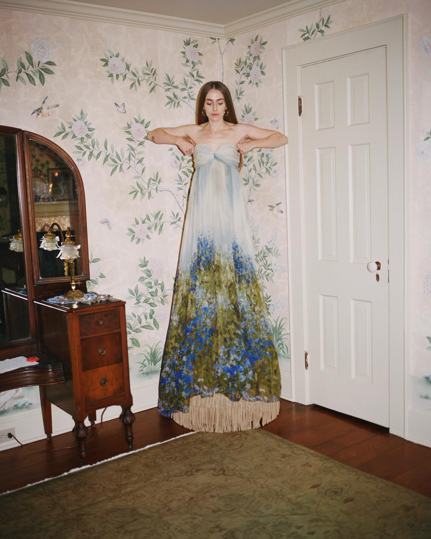 A woman in a flowing strapless dress poses in an indoor setting with floral wallpaper, a wooden dresser, and a brown rug.