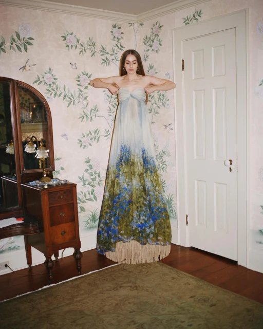 A woman in a flowing strapless dress poses in an indoor setting with floral wallpaper, a wooden dresser, and a brown rug.