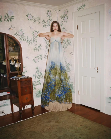 A woman in a flowing strapless dress poses in an indoor setting with floral wallpaper, a wooden dresser, and a brown rug.