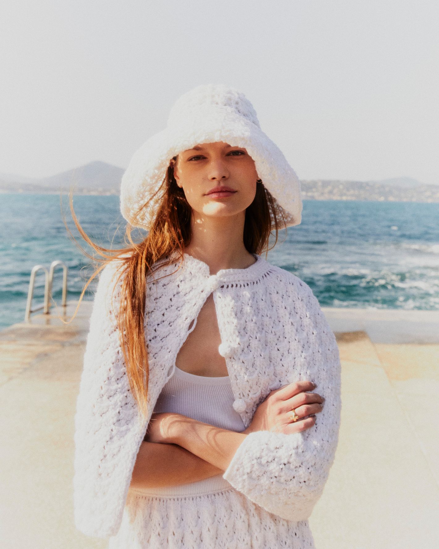A woman in a white crochet outfit and sunhat stands on a light-colored concrete platform with a view of the ocean and distant land.