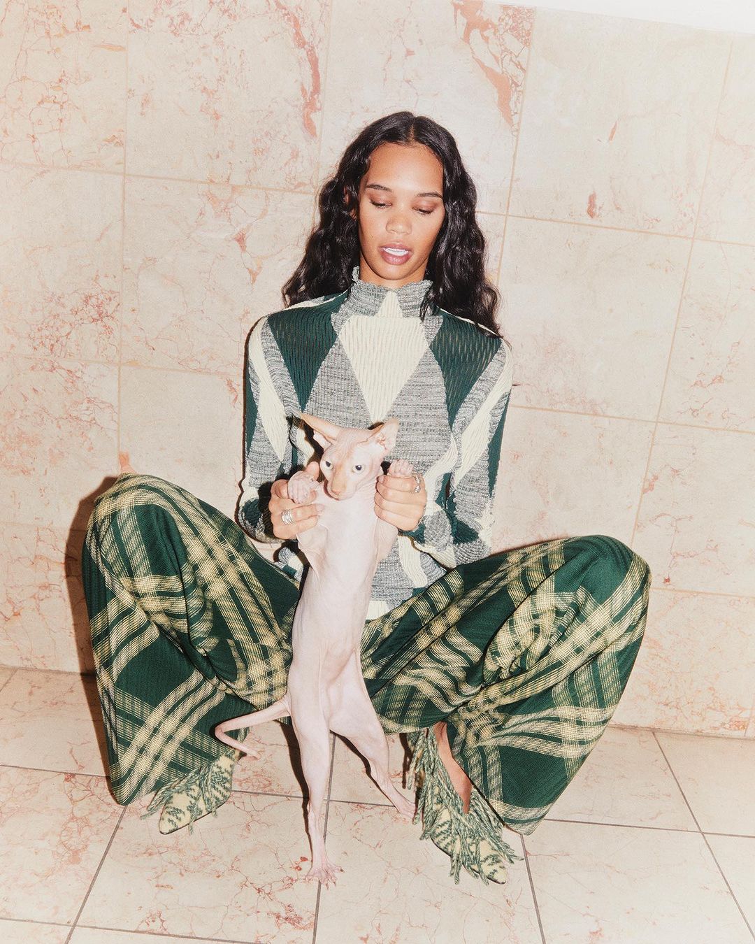 Woman poses indoors holding a hairless cat. She wears a patterned top and plaid pants, with a tile backdrop and floor.