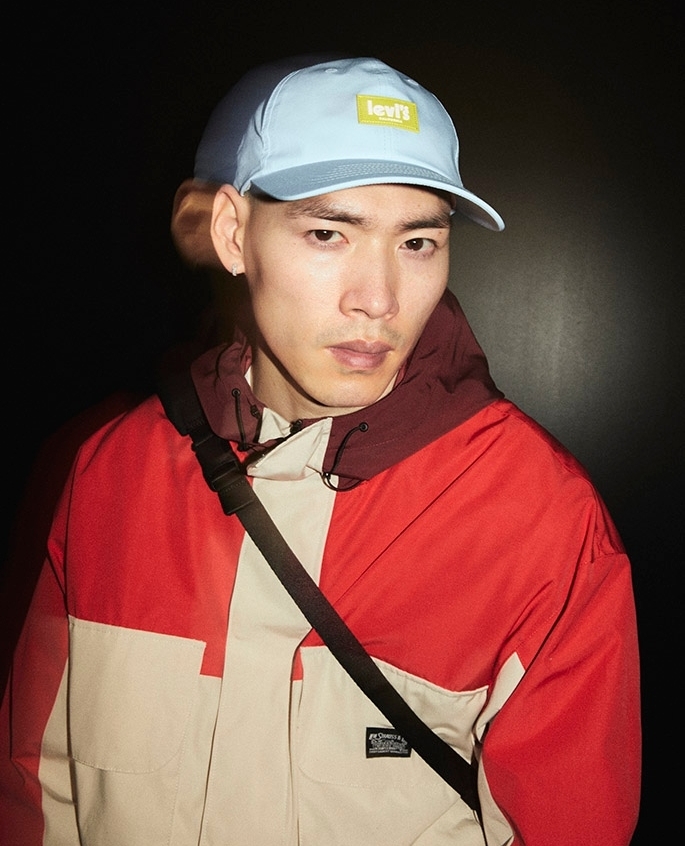 Portrait of an Asian man wearing a Levi's cap and color-blocked hooded jacket, posing against a dark background, studio lighting.
