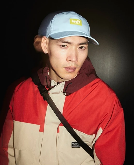 Portrait of an Asian man wearing a Levi's cap and color-blocked hooded jacket, posing against a dark background, studio lighting.