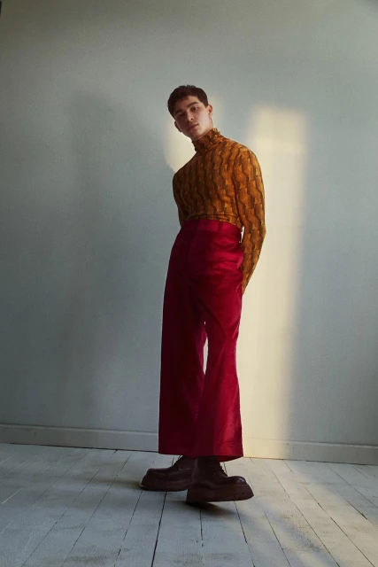 Stylish young man in a patterned turtleneck and red trousers posing in a photo studio with soft lighting.