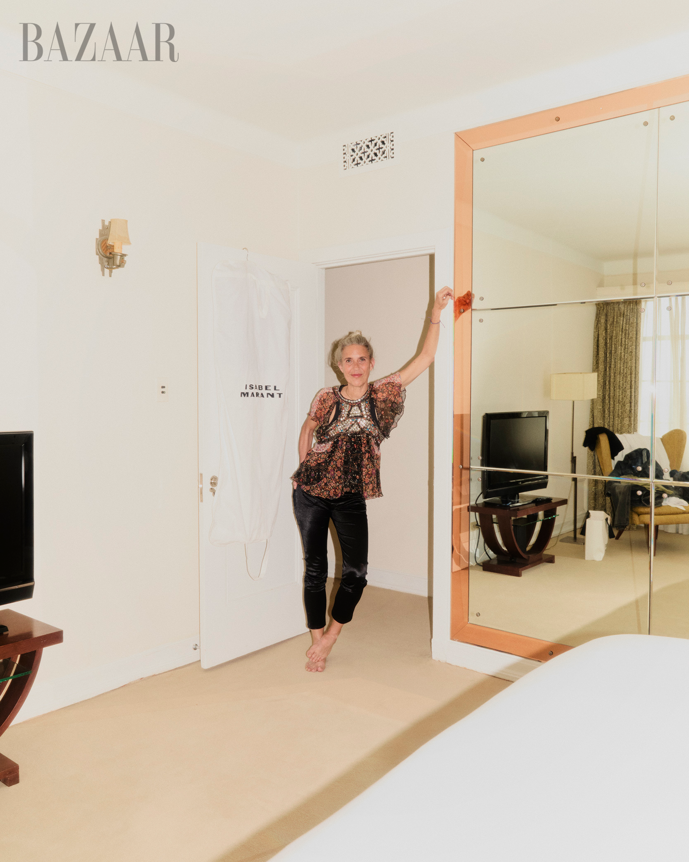 Blonde woman in patterned top and velvet pants poses barefoot in a light room with a mirror and white door, inside a Harper's Bazaar photo.