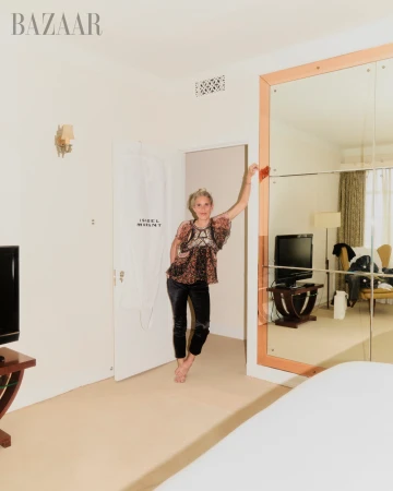 Blonde woman in patterned top and velvet pants poses barefoot in a light room with a mirror and white door, inside a Harper's Bazaar photo.