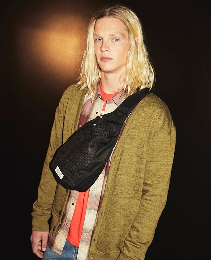 An adult male with blonde hair, wearing a cardigan and plaid shirt, poses in front of a dark background with a black shoulder bag.