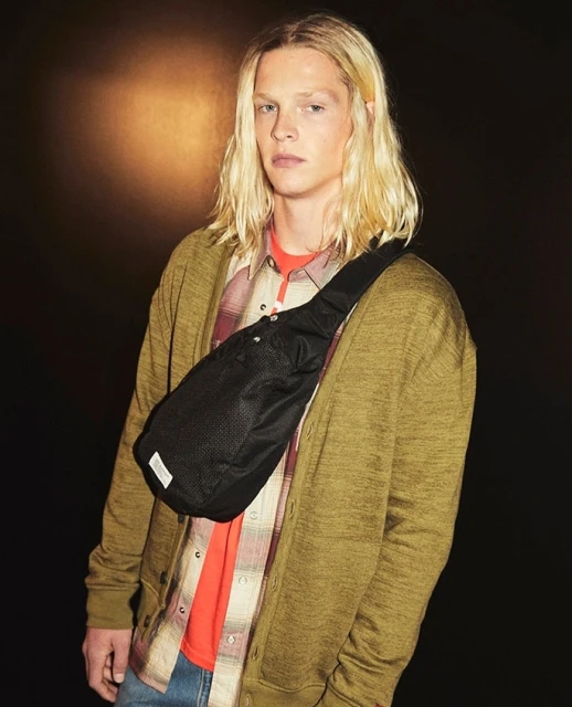 An adult male with blonde hair, wearing a cardigan and plaid shirt, poses in front of a dark background with a black shoulder bag.