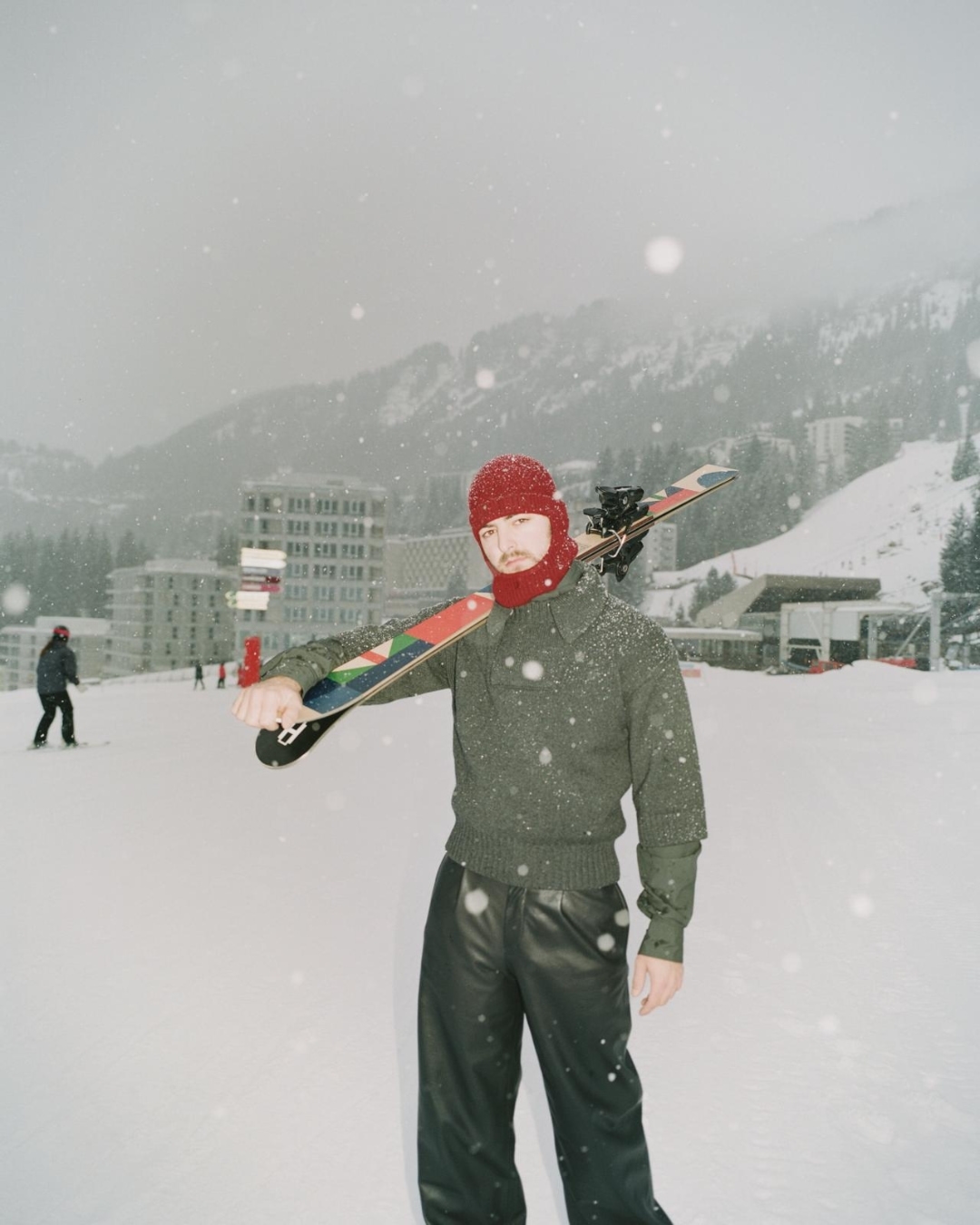 A person in a red balaclava and green sweater stands in the snow on a ski slope with skis over their shoulder. Snowy mountains in the distance.