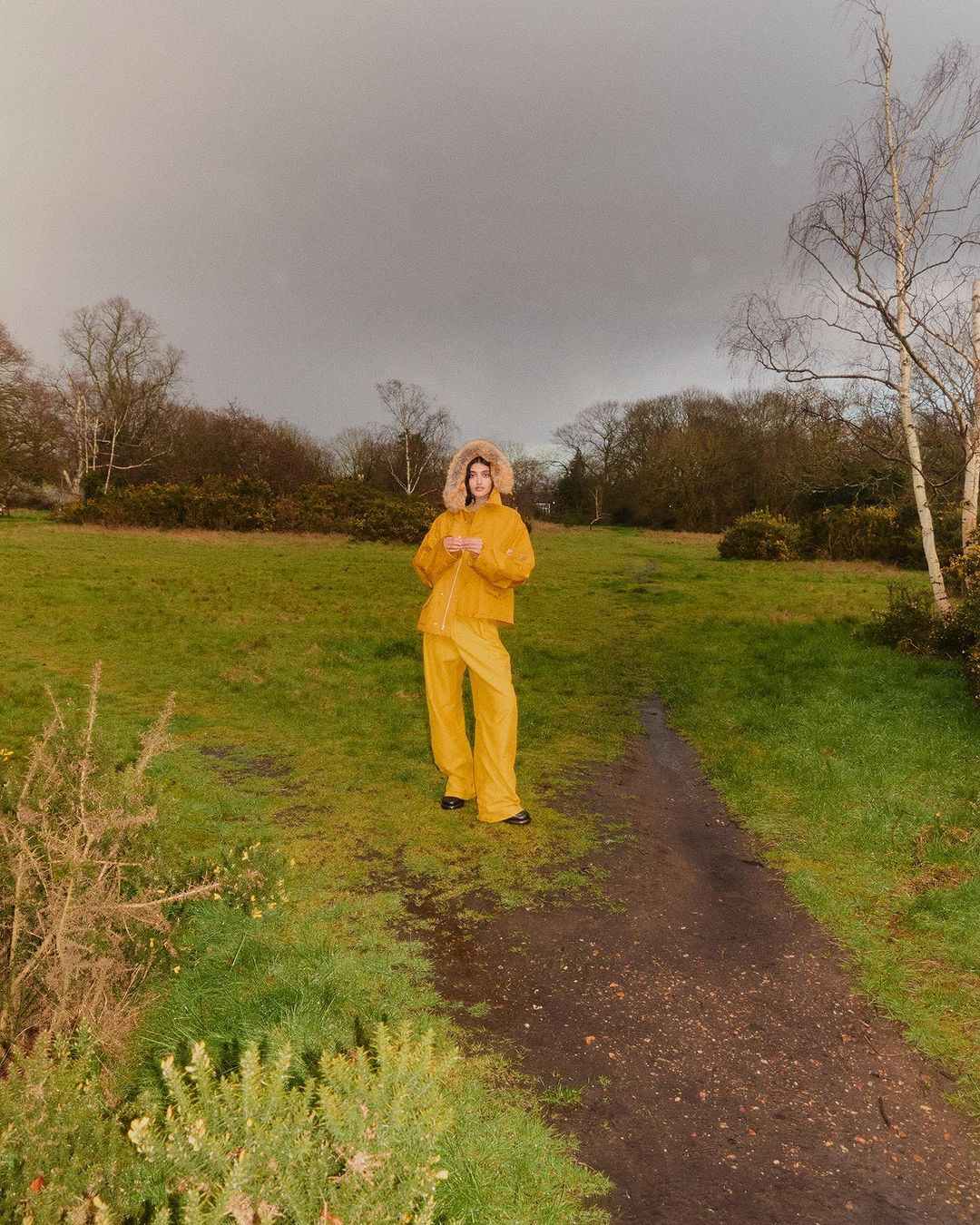 Model wearing a bright yellow hooded jacket and matching trousers standing on a dirt path in a grassy field under an overcast sky.