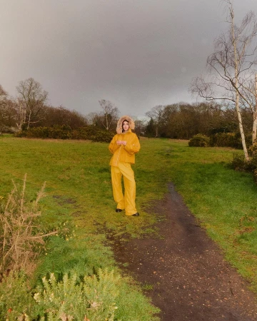 Model wearing a bright yellow hooded jacket and matching trousers standing on a dirt path in a grassy field under an overcast sky.
