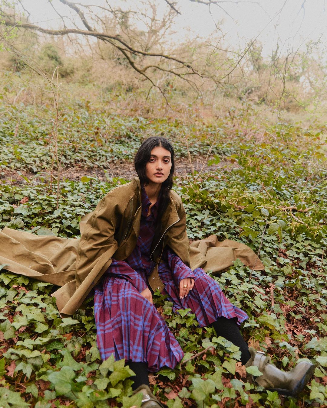 Woman sits among ivy in an olive coat and plaid dress, gazing at the camera. Outdoor fashion shot with natural light.
