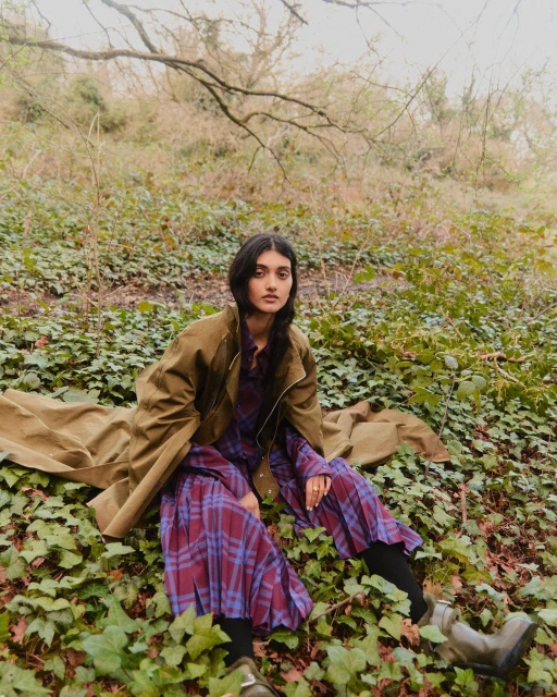Woman sits among ivy in an olive coat and plaid dress, gazing at the camera. Outdoor fashion shot with natural light.