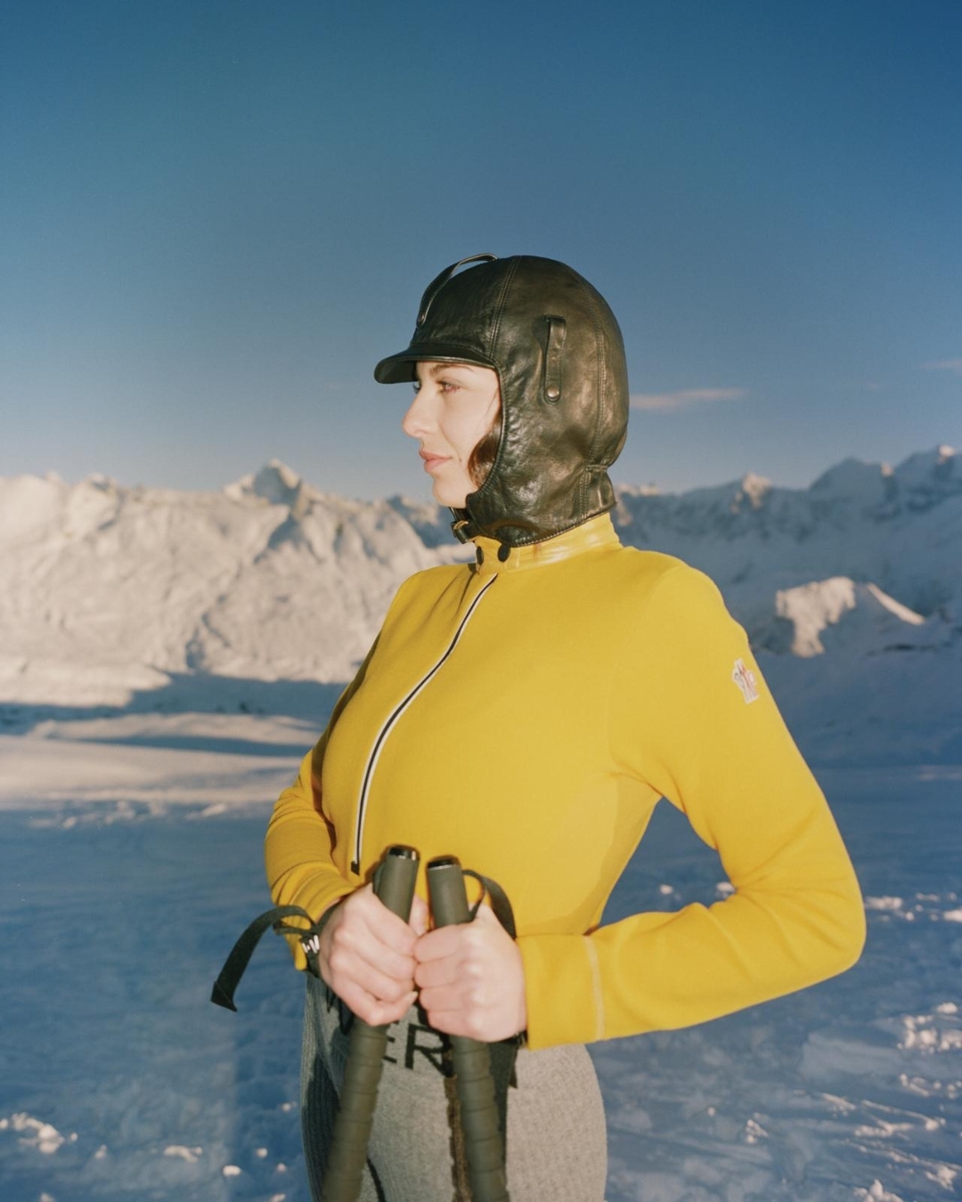 Woman in yellow ski suit and leather helmet standing in snowy mountains. Ski poles in hand, looking away from the camera on a bright day.