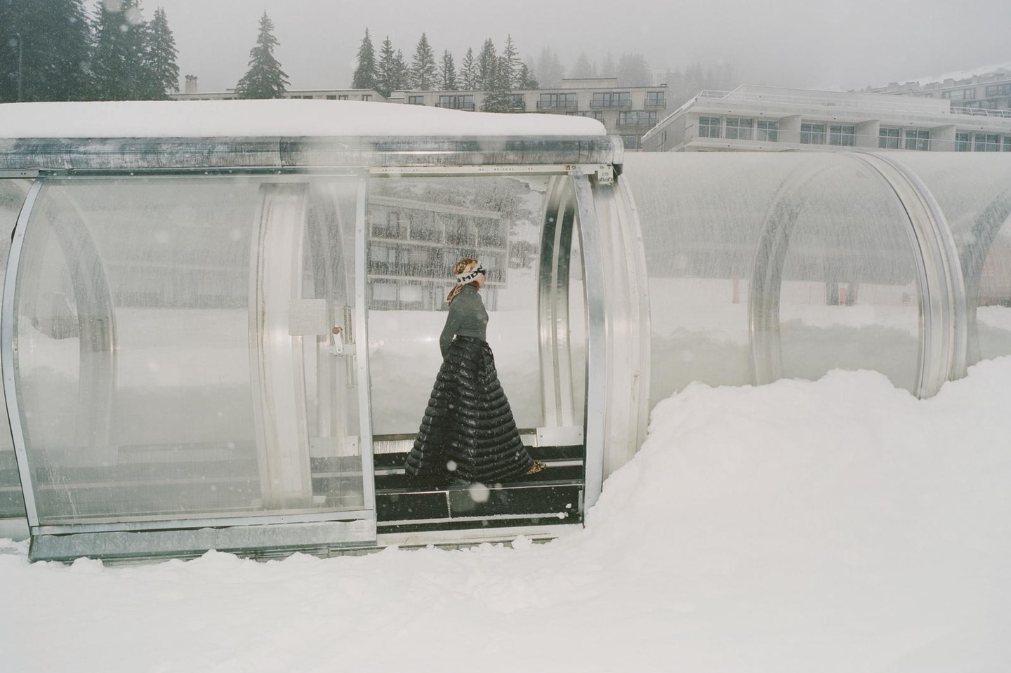 Woman in a long quilted skirt walks through a snow-covered glass tunnel on a winter day, surrounded by snow-laden trees and buildings.