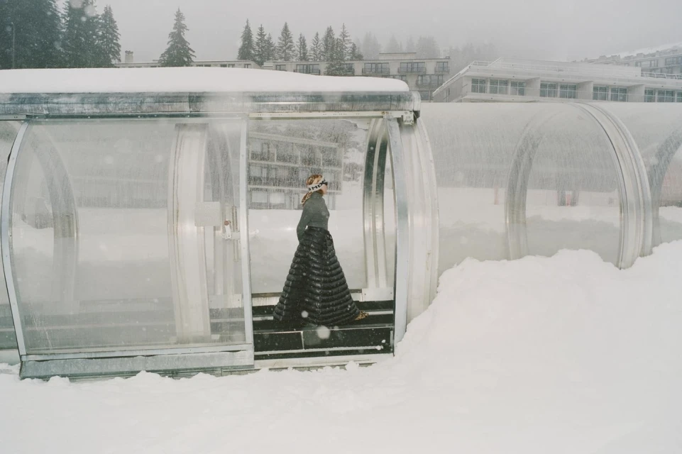 Woman in a long quilted skirt walks through a snow-covered glass tunnel on a winter day, surrounded by snow-laden trees and buildings.