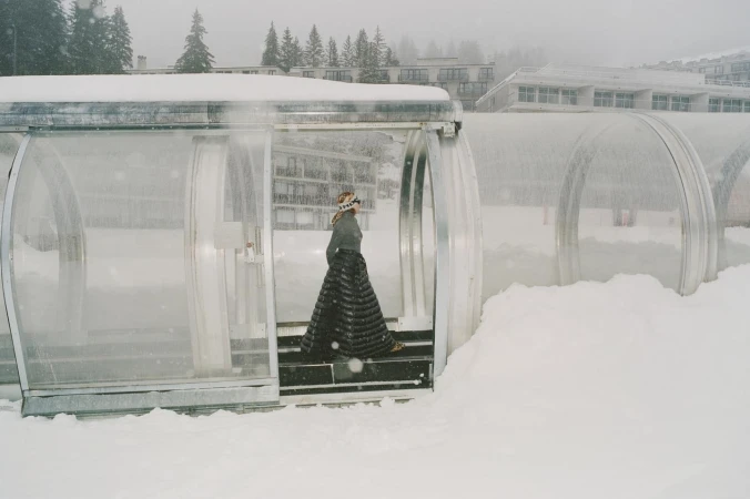Woman in a long quilted skirt walks through a snow-covered glass tunnel on a winter day, surrounded by snow-laden trees and buildings.