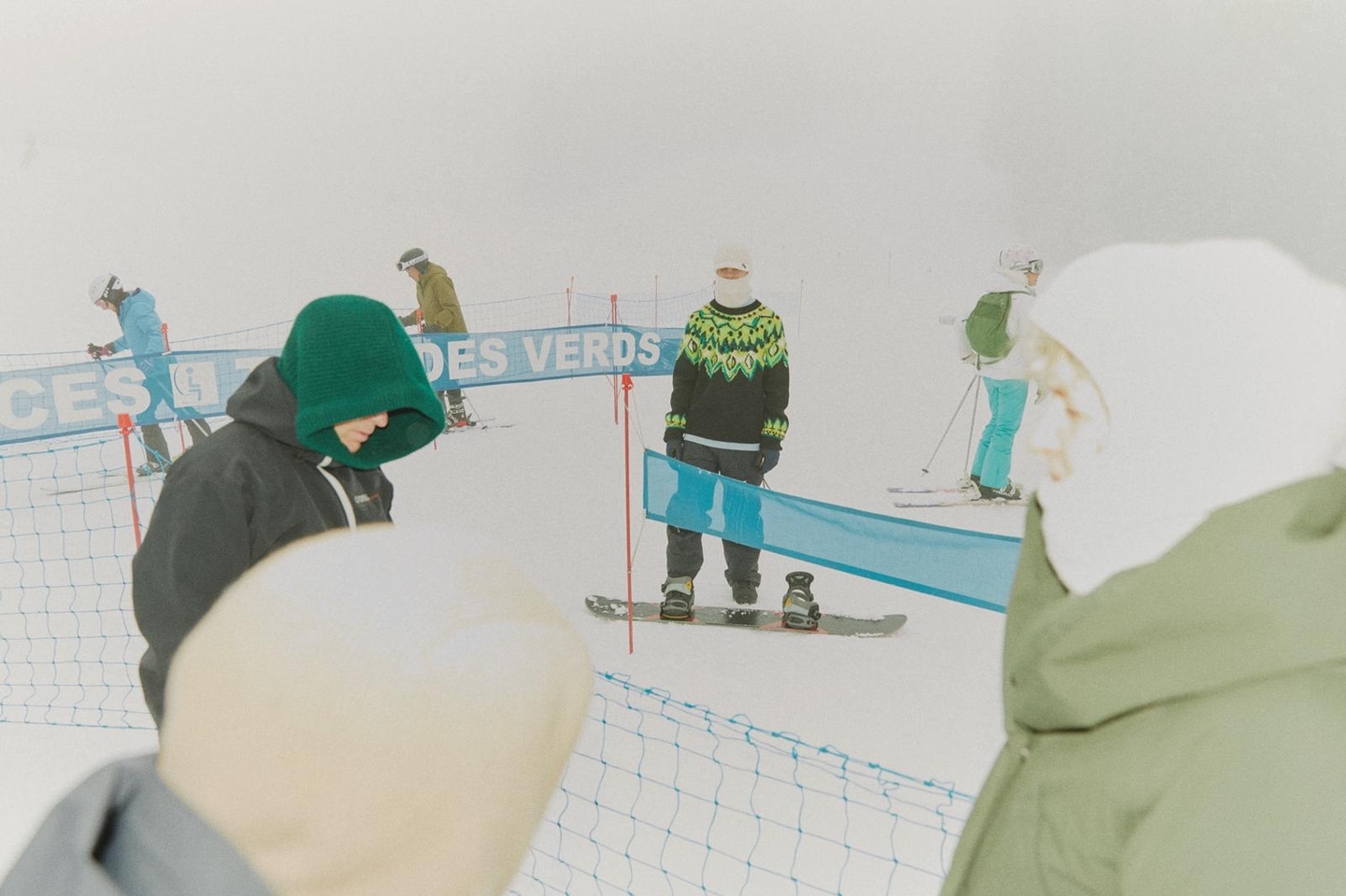 Several skiers and snowboarders are on a snow-covered mountain with a slight fog and blue safety nets in the background.