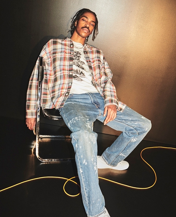 Young man with braids seated on a metal chair wearing a plaid shirt and paint-splattered jeans against a dark background