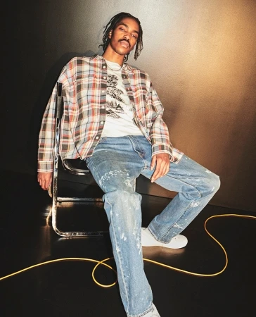 Young man with braids seated on a metal chair wearing a plaid shirt and paint-splattered jeans against a dark background