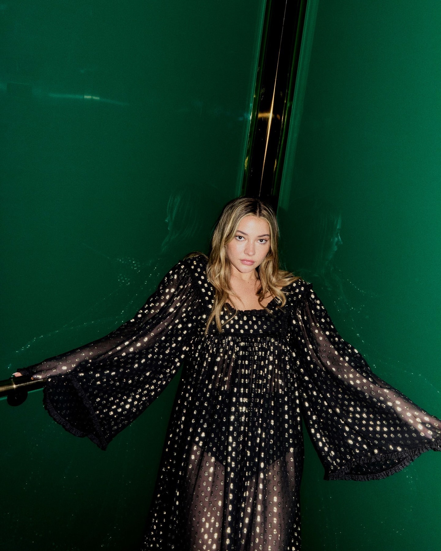 A woman with wavy blond hair poses against a green backdrop, wearing a black dotted dress with wide sleeves and a square neckline.