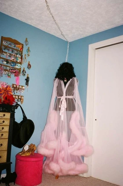 Pink faux fur robe with white trim on mannequin, accessorized with jewelry, gold shoes, and black purse against blue wall.