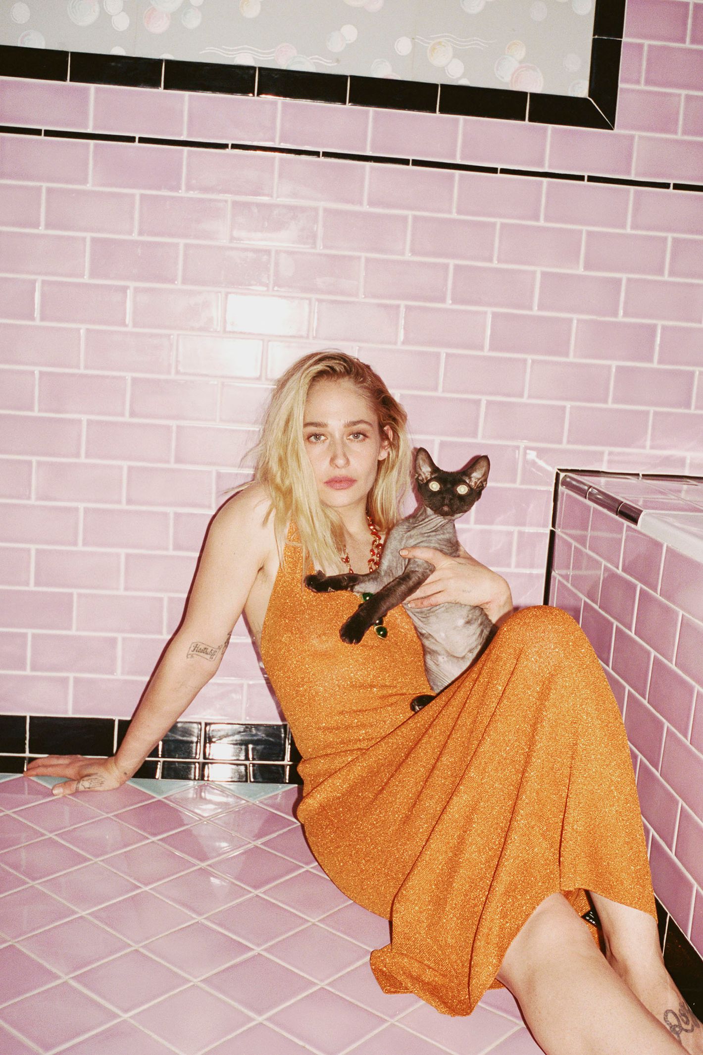 Woman with blonde hair sits on a pink-tiled floor, wearing an orange dress and holding a gray cat, posing against a pink-tiled wall.