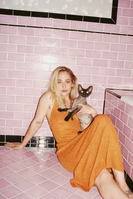 Woman with blonde hair sits on a pink-tiled floor, wearing an orange dress and holding a gray cat, posing against a pink-tiled wall.