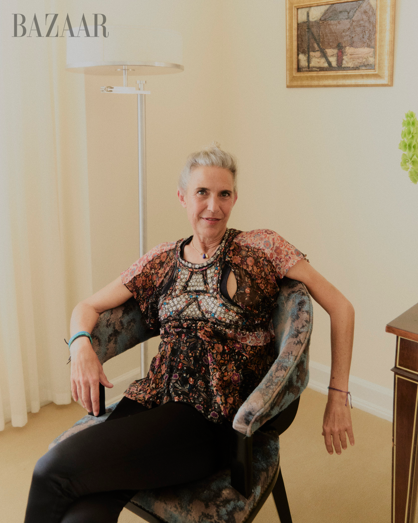 Older woman with short grey hair sits in blue chair in an indoor setting with painting on the wall in the background.