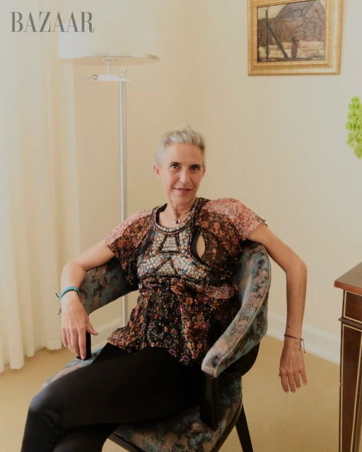 Older woman with short grey hair sits in blue chair in an indoor setting with painting on the wall in the background.