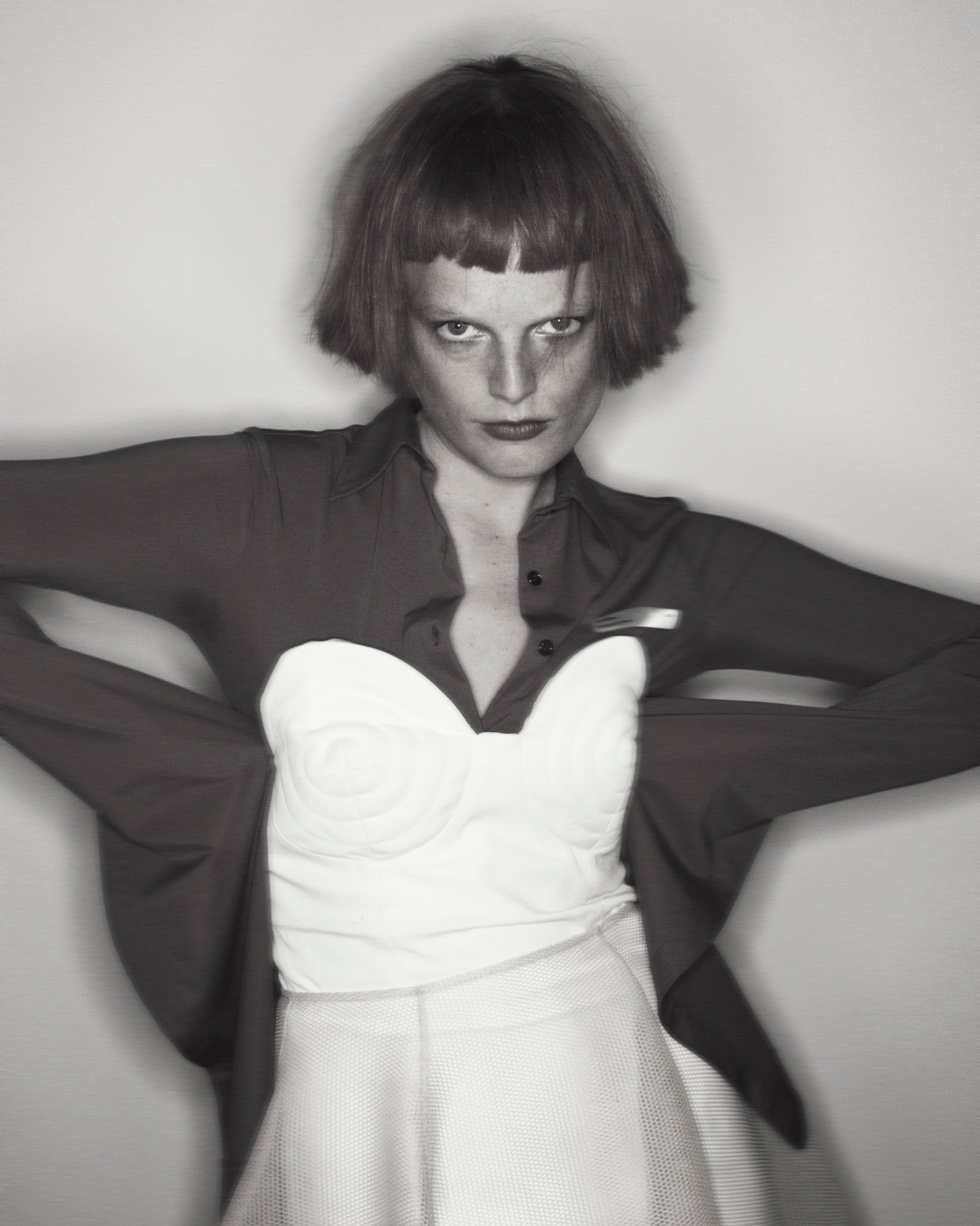 Monochromatic portrait of a person with a red bob cut wearing a white bustier, red shirt, and white skirt against a gray background.