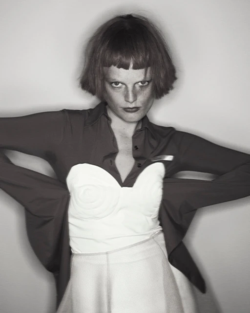Monochromatic portrait of a person with a red bob cut wearing a white bustier, red shirt, and white skirt against a gray background.