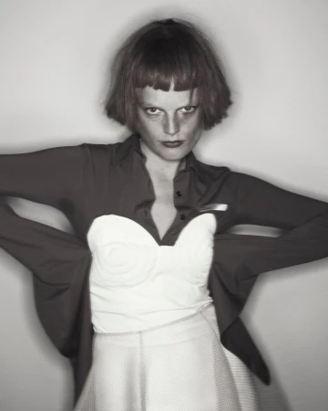 Monochromatic portrait of a person with a red bob cut wearing a white bustier, red shirt, and white skirt against a gray background.