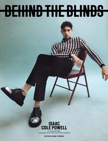 A young adult man in a patterned sweater and black pants sits on a red folding chair against a blue background for a fashion editorial.