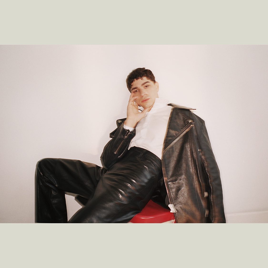 Portrait of a young adult male in a white shirt and black leather pants, seated on a red stool with a jacket draped over his shoulder.