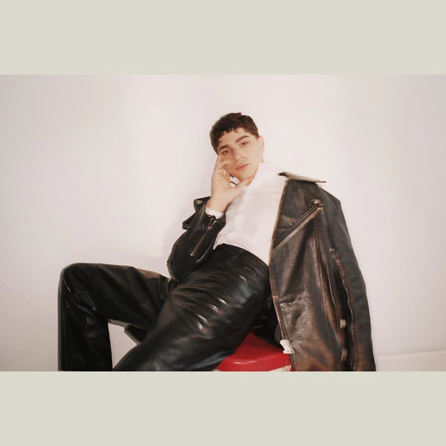 Portrait of a young adult male in a white shirt and black leather pants, seated on a red stool with a jacket draped over his shoulder.