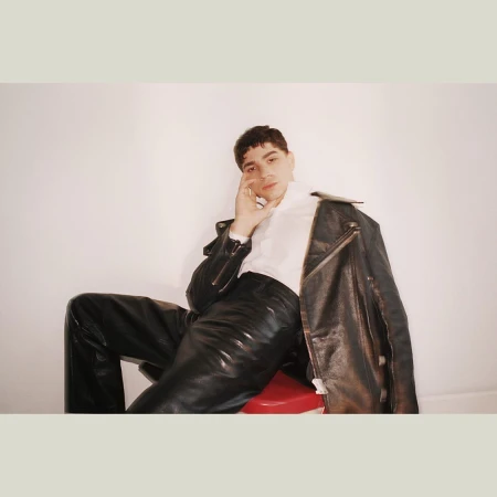 Portrait of a young adult male in a white shirt and black leather pants, seated on a red stool with a jacket draped over his shoulder.