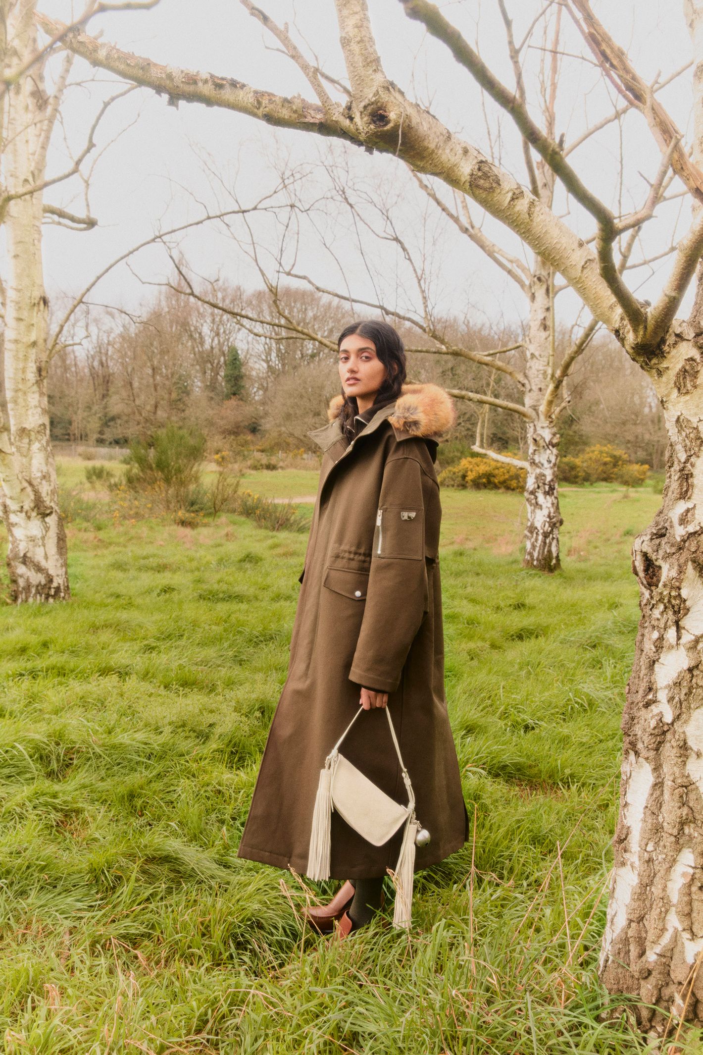 A woman in a fur-trimmed hooded coat stands in a grassy field, holding a handbag, with trees in the background during daylight hours.