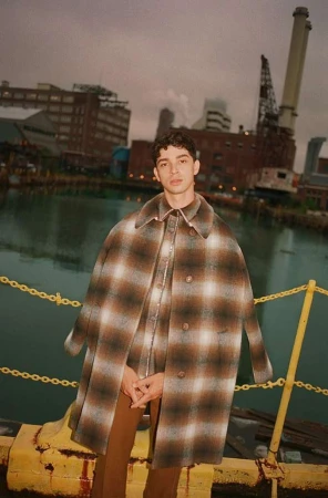Male fashion model poses in a brown plaid coat and pants, standing outside near water and industrial buildings on an overcast day.